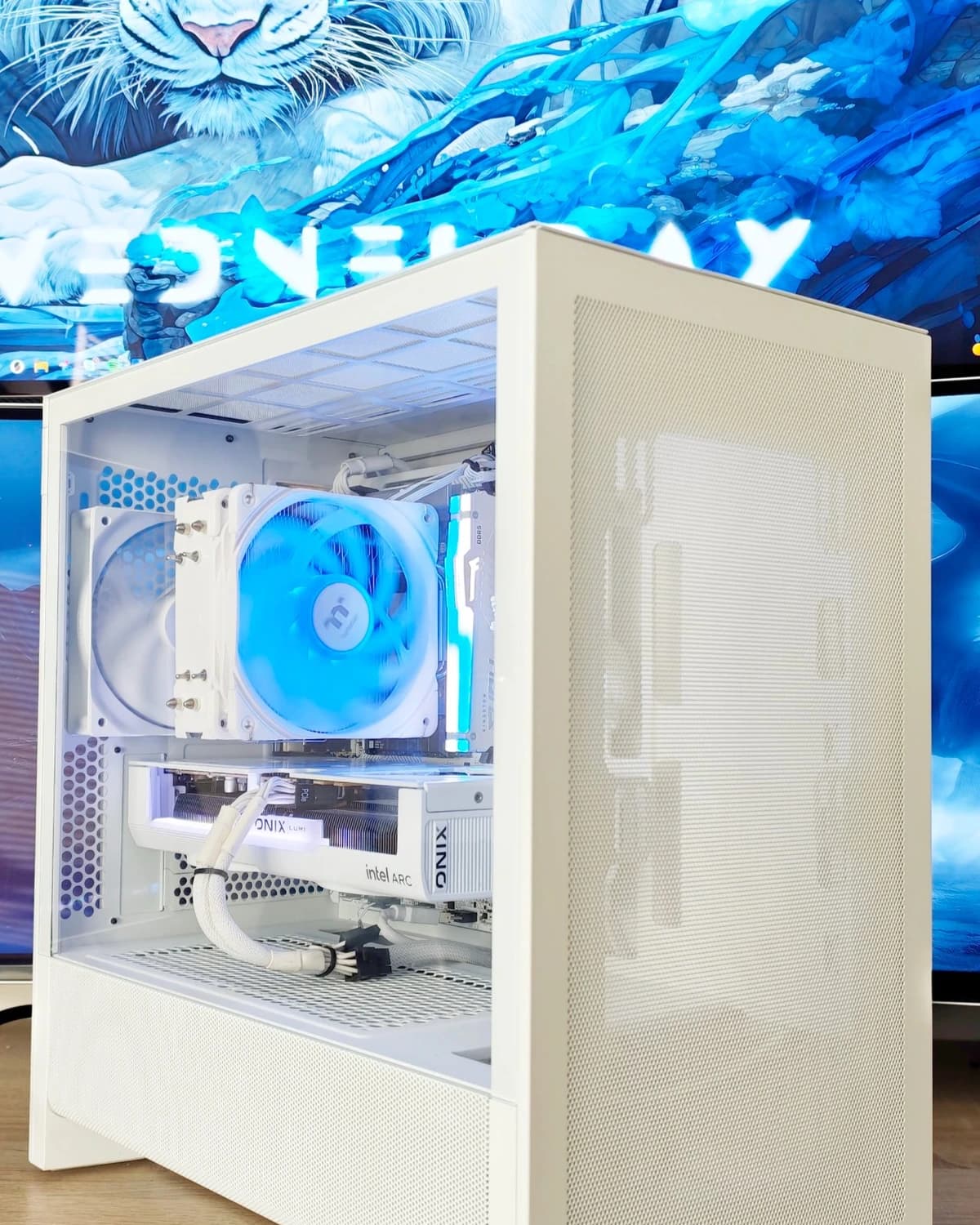 White NZXT H3 Flow gaming PC with a blue-lit cooler and an Intel Arc B580 graphics card in front of a monitor showing a white tiger