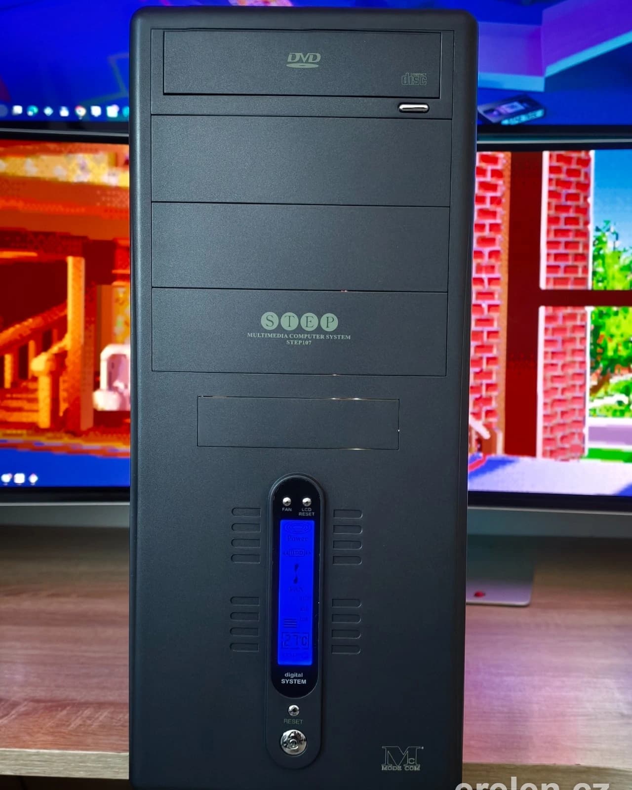 Retro počítačová skříň STEP s DVD mechanikou a modrým LCD displejem před monitorem s pixelartovou hrou
